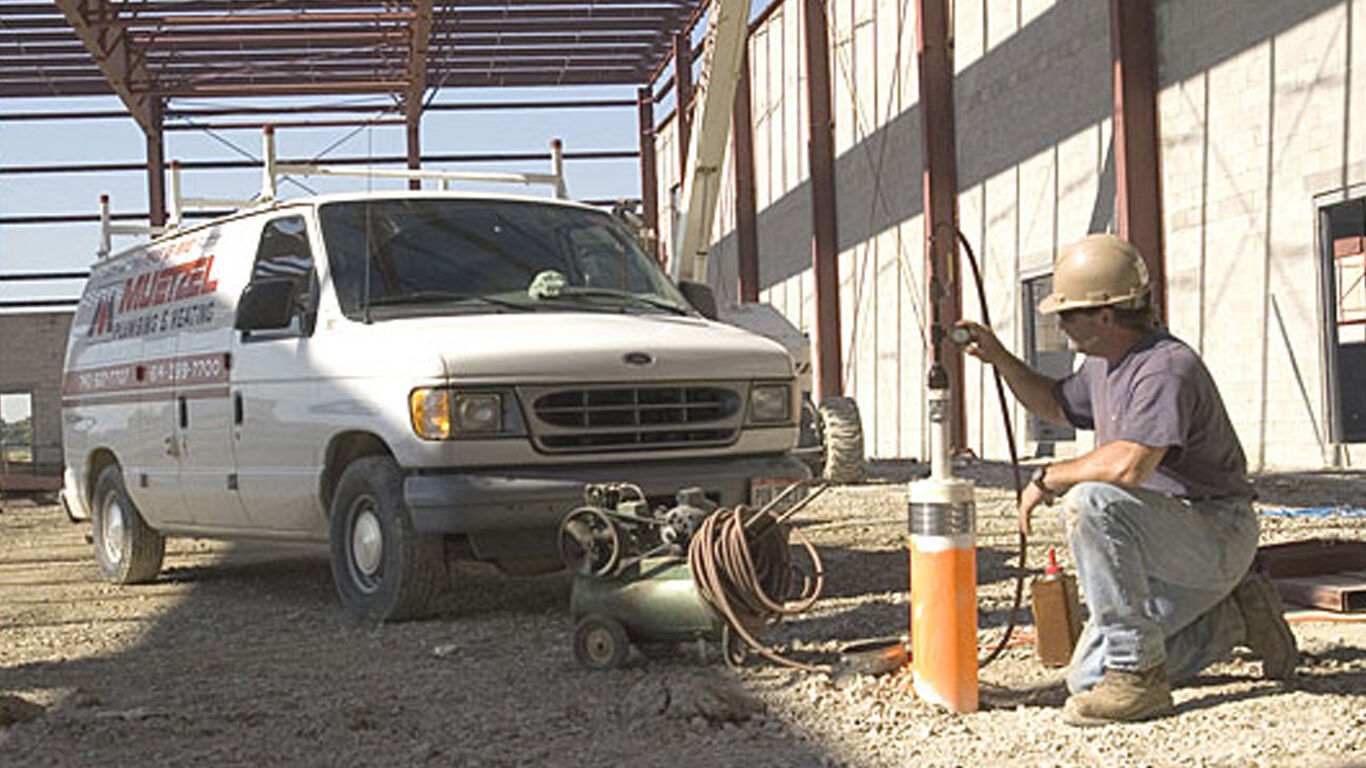 A man on one knee using an air compressor to blow air into pipe on a job site